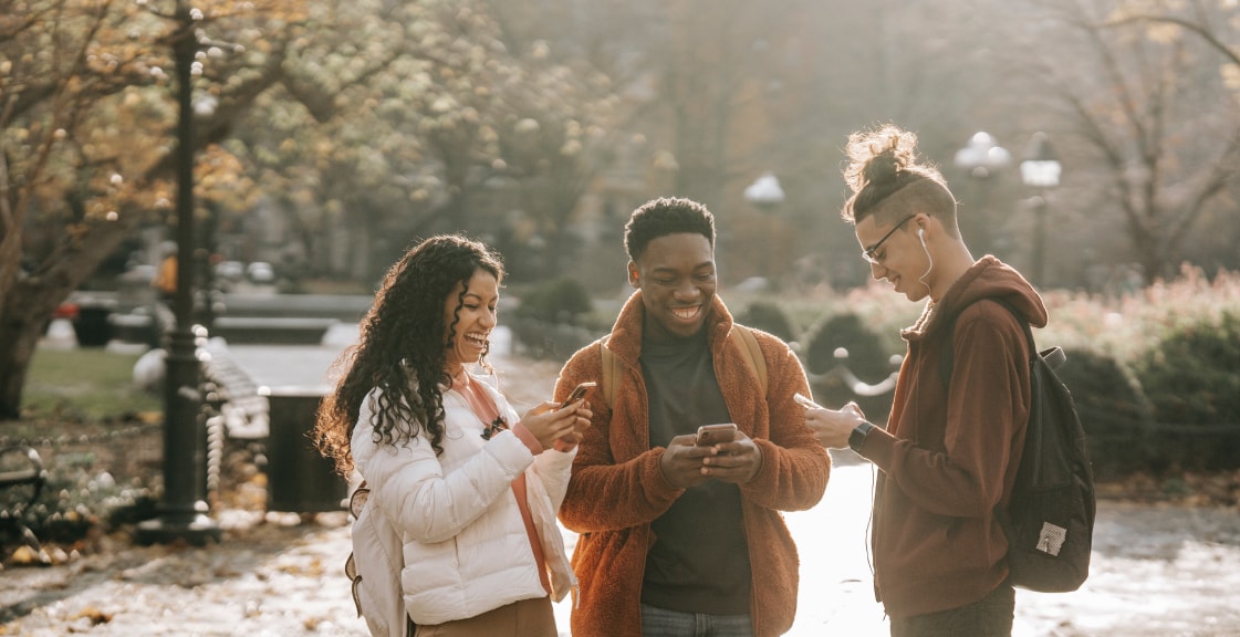Three friends standing together at a park holding their phones and looking down on their screens while smiling.